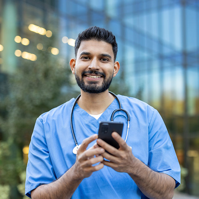 A smiling healthcare professional in scrubs uses a smartphone outside a modern building. His stethoscope is around his neck.