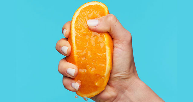 Indoor photo of hand holding orange and clenching a fists while squeezing juice over blue background