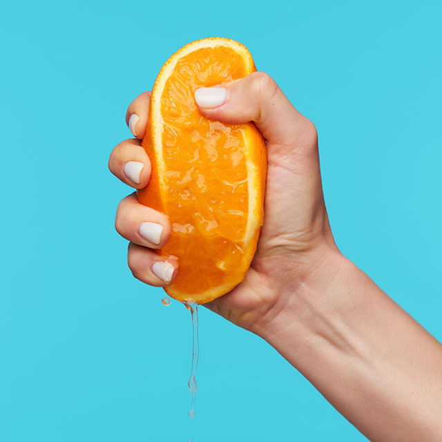 Indoor photo of hand holding orange and clenching a fists while squeezing juice over blue background