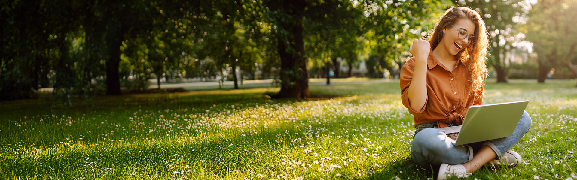 Happy woman with laptop working in a sunny park on a green lawn.